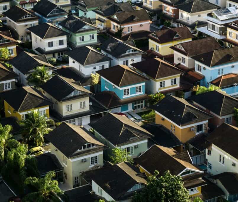 An aerial view of a densely packed residential neighborhood featuring colorful two-story houses with dark gabled roofs and tropical palm trees.