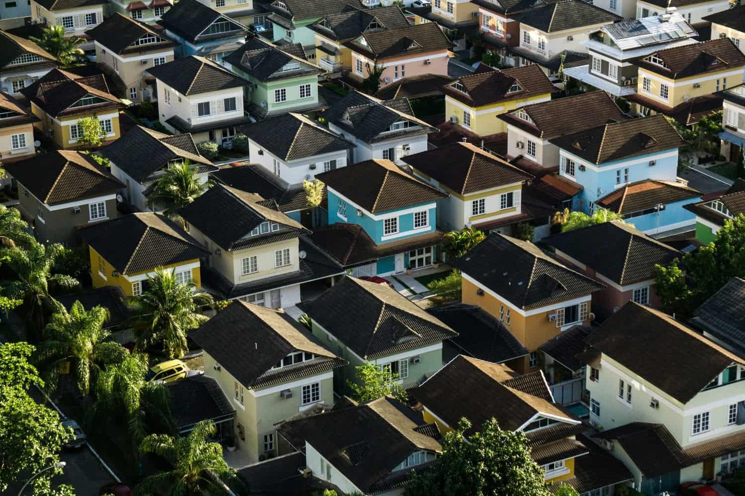 An aerial view of a densely packed residential neighborhood featuring colorful two-story houses with dark gabled roofs and tropical palm trees.